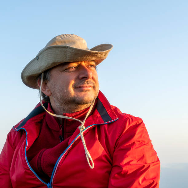 A mature man is looking away and he is wearing a hat. He is against blue sky at sunset time.