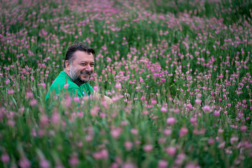 A man is sitting in the pink flowers garden. He is looking at the objective and  smiling.