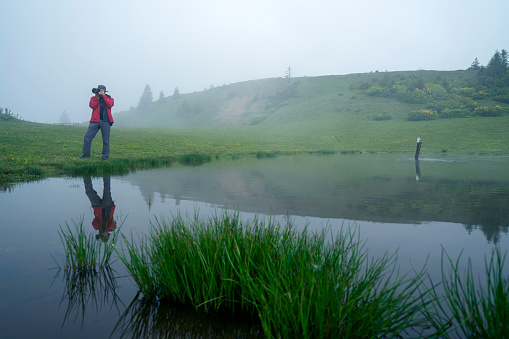 A photographer is shooting photography by the lake in the region of Black Sea. The weather is foggy and there is reflection on the lake.