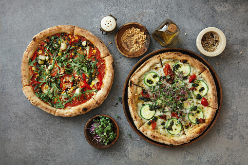 Vegetarian pizza with tomato sauce, cauliflower, corn, arugula, nuts and microgreen. Vegetarian pizza with zucchini, sun-dried tomatoes source pesto, green sprouts and nuts. Flat lay top-down composition on concrete background.