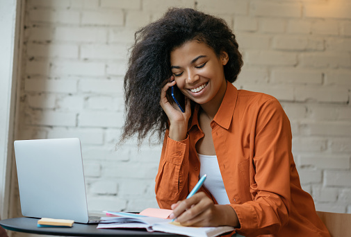 Young smiling businesswoman using laptop, talking on mobile phone, taking notes, working startup project in modern office. Successful business concept. Portrait of beautiful woman freelancer working at home