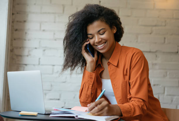 mulher de negócios sorridente usando laptop, falando no celular, tomando notas, trabalhando projeto de startup em escritório moderno. conceito de negócio de sucesso. retrato de mulher bonita freelancer trabalhando em casa - agenda de telefones - fotografias e filmes do acervo