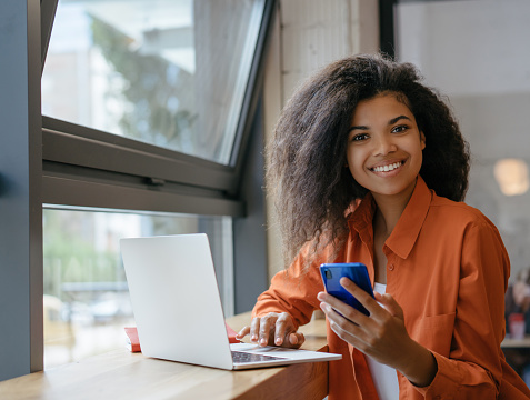 Young beautiful woman working in office. Portrait of smiling African American businesswoman using laptop computer and internet, holding mobile phone. Successful business concept