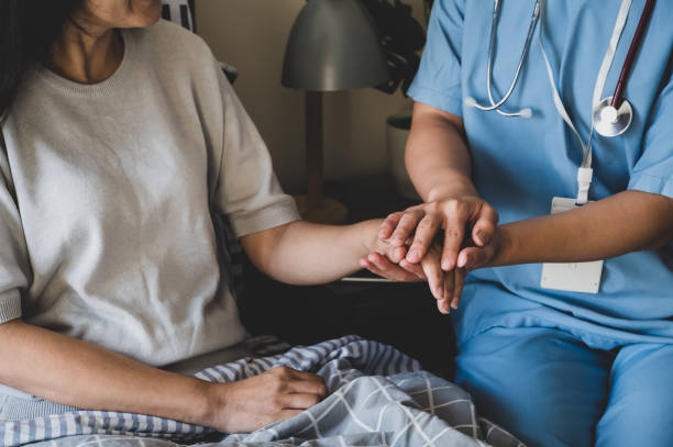 Close-up of a female nurse holding hands for reassuring her female patient on bed. Female home caregiver giving thumbs-up and moral support to an elderly female patient for her speedy recovery from her illness in bedroom. simple living stock pictures, royalty-free photos & images