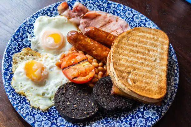 Plate of Scottish breakfast with two fried eggs, bacon, two sausages, two black puddings, baked beans, potato bread, tomato and two slices of toast.