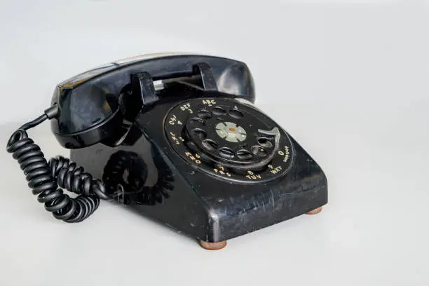 An old black rotary dial phone. It is well used, slightly scratched and shows spots of dirt. Whitish background, shallow depth of field.