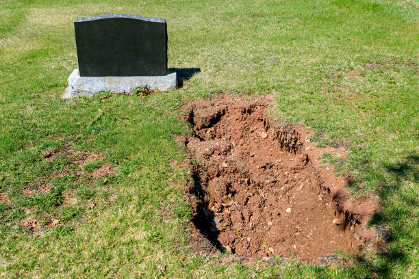 A fresh grave by a blank black tombstone. The hole is about half filled with dirt. Green grass around stone and hole, sunny day.
