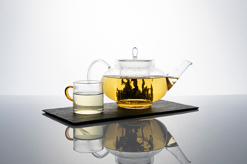 Transparent teapot and cup with brewed green herbal tea on white background.