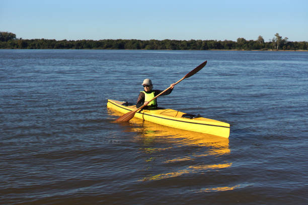 A man leisurely paddles a bright orange kayak on a serene river. The calm water reflects the dense greenery of the surrounding forest, creating a peaceful setting ideal for outdoor water sports and relaxation.