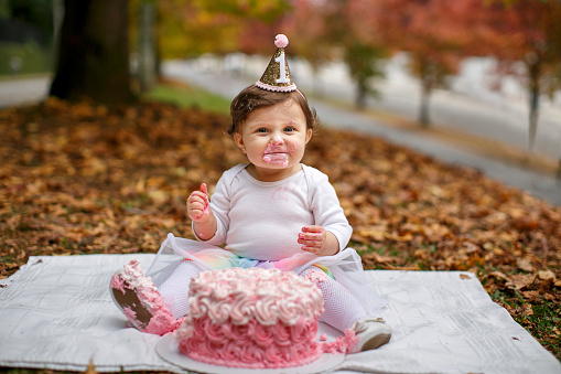 Curitiba, Brazil - April 29, 2021: Baby girl sitting on the ground behind a pink cake and wearing a paper hat celebrating her first anniversary.