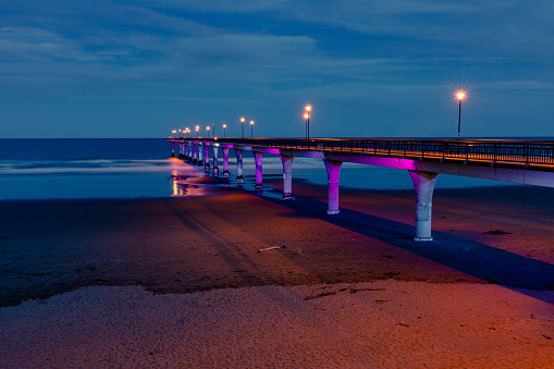 This November 2020 long-exposure photo shows the New Brighton Pier in Ōtautahi Christchurch, Aotearoa New Zealand at dusk. The Pacific Ocean is at low tide, exposing the pillars of the pier. The pillars are illuminated by coloured LED lights. The blurred figures of multiple people are seen on the pier.