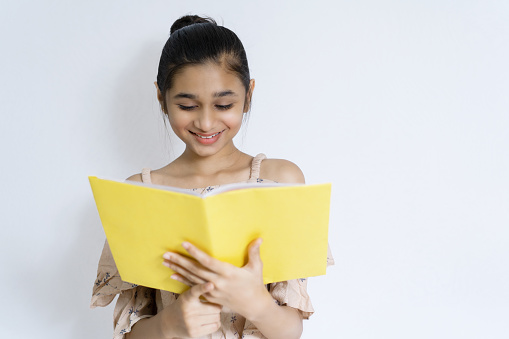 Happy Asian, Indian happy teenager girl student standing against an isolated white background and reading a book with a smile.