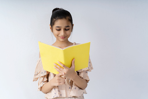 Happy Asian, Indian happy teenager girl student standing against an isolated white background and reading a book with a smile.