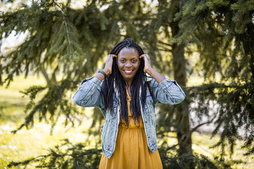 Portrait of young smiling african woman standing in park and fixing hair while looking at camera