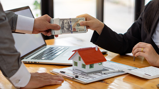 A real estate agent introduces clients to a home layout with contract documents and a laptop and dollar bill on the table. Customers interested in purchasing a home sign their signature to complete a home purchase contract.