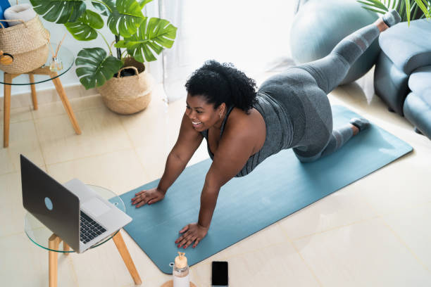 Young African curvy woman doing pilates virtual fitness class with laptop at home - Sport wellness people lifestyle concept Young African curvy woman doing pilates virtual fitness class with laptop at home - Sport wellness people lifestyle concept exercise class photos stock pictures, royalty-free photos & images