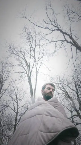 Young man with beard covered with gray blanket in the forest in winter