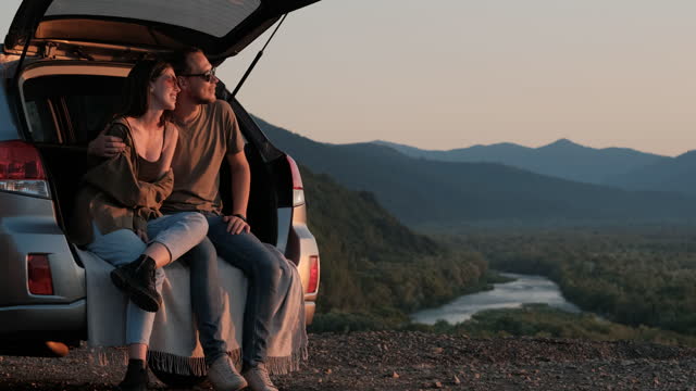 Young Traveler Couple on a Road Trip, Man and Woman Sitting on the Opened Trunk of Their Suv Car, Mountains and River Canyon on the Background
