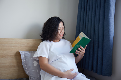 A young Vietnamese pregnant woman with short hair lying on a bed while reading a book