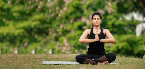 Outdoor image of Young Asian/ Indian woman sitting in Namaste or prayer yoga position and relaxing at park. Concept of Yoga and healthy lifestyle.