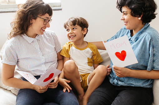 Boy with his two mothers holding greeting cards. Smiling women looking at their son while sitting on sofa with mothers day card.