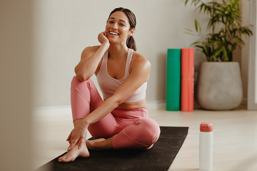 Female in sportswear sitting on yoga mat and smiling after workout. Woman taking a break from workout session at gym.