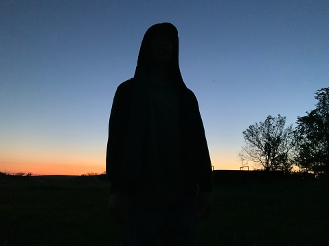 Silhouette of a person center frame in a hoodie overlooking a field. The sky is blue with just a rim of sunlight making a rainbow of cloud reflections across the most falling sky. A far off springtime tree is outlined in the corner.
