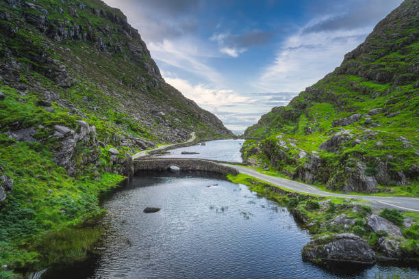 winding narrow road crossing stone wishing bridge in gap of dunloe, black valley - winding road sunlight field cultivated land imagens e fotografias de stock