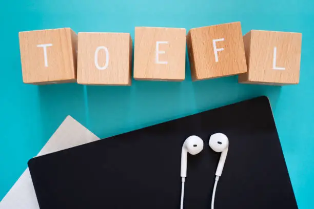 TOEFL listening; Five wooden blocks with TOEFL text of concept, a black notebook, a piece of paper, and a pair of earphones.