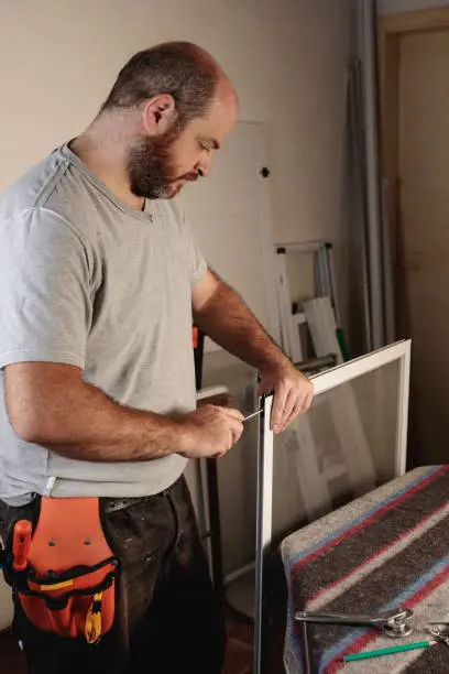 Photo of man working in an aluminum carpentry shop, assembling an aluminum window screen. Vertical image.