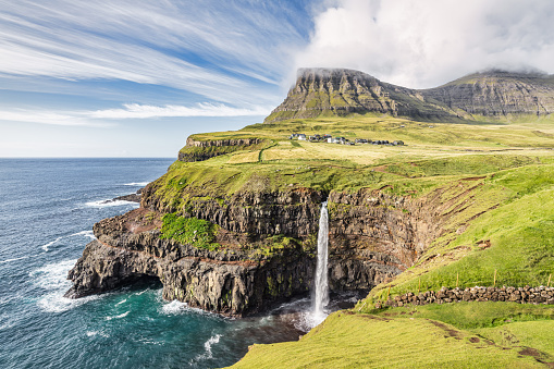 Faroe Islands beautiful iconic natural Mulafossur - Múlafossur Waterfall with Gasadalur Village in the background under blue sunny summer sky. Mulafossur Waterfall, Gasadalur, Vagar Island, Faroe Islands, Denmark, Nordic Countries, Europe