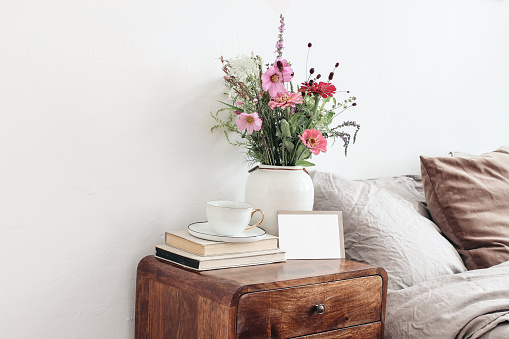 Cup of coffee and books on retro wooden bedside table. Blank greeting card mockup. White ceramic vase. Buquet of cosmos and zinnia flowers, beige linen, velvet pillows in bed. Scandinavian interior.
