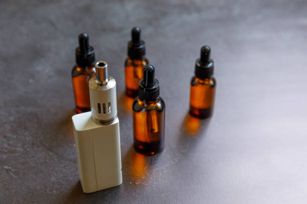 istock Vaping device e-cigarette electronic cigarette and liquid bottles on dark black stone shale background. Vape device for alternative smoking. Vaping shop concept. Gadget for vaper. Vaping accessories 1315989161