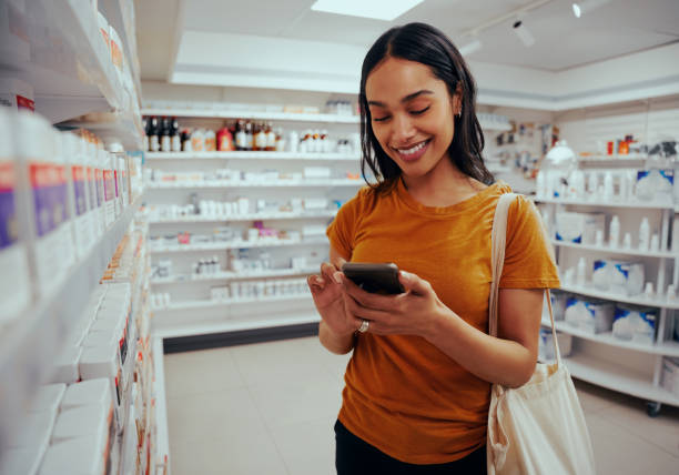 Young woman smiling while using smartphone standing against shelf in pharmacy