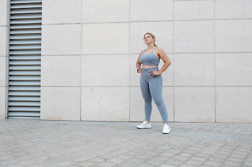 Pretty fit young plus size woman in sports bra and leggings standing outdoors after jogging