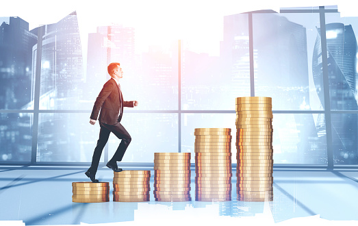 Side view of young businessman climbing stacks of coins in blurry panoramic office. Concept of financial success and career growth. toned image double exposure