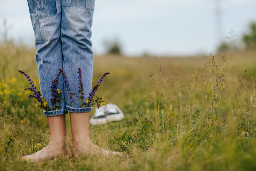 Close-up young female legs standing on green spring grass with wildflowers in legs of denim trousers
