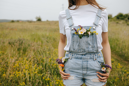 One young woman in white t-shirt and suspenders  jeans with wildflowers in pocket standing on blooming meadow, enjoying sunny day in springtime nature