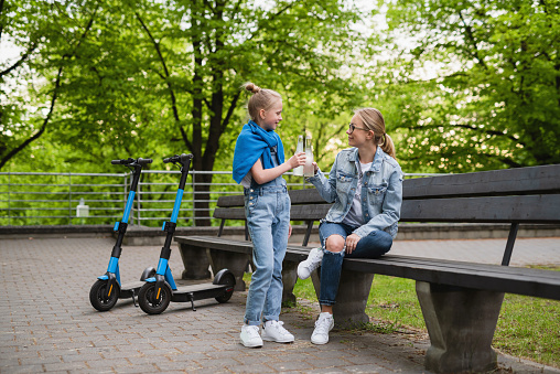 Happy mother and daughter with electric scooters are drinking lemonade in city park