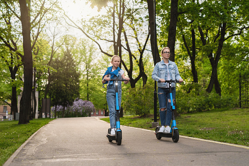 Happy mother and daughter riding electric scooters  in city park