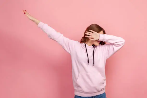 Photo of Portrait of young female in hoodie raising hands in dab dance pose, celebrating success, showing gesture of triumph, popular internet move