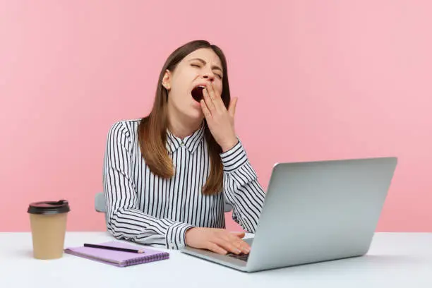 Tired overworked woman office worker yawning sitting at workplace with laptop, exhausted with job routine, showing disinterest. Indoor studio shot isolated on pink background