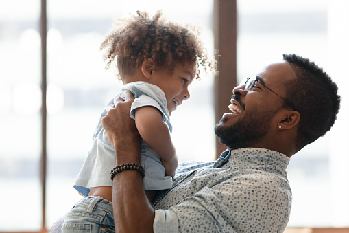 Happy daddy holding kid in arms, hugging toddler boy. African American dad enjoying being father, spending time and playing with cute preschooler son, talking to child, laughing. Parent concept