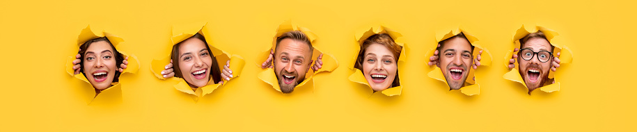 Group of happy people smiling and looking at camera while ripping bright yellow paper and peeking out from holes