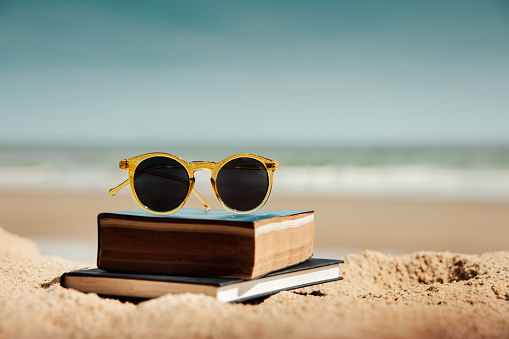 Reading Book Outdoor in Summer Concept. Book and Sunglasses on the Beach Sand in front of the Sea on Sunny Day. Lifestyle on Vacation