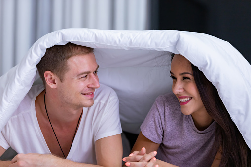 Portrait of young happy playful couple relaxing in comfortable cozy bed, cheerful smiling man and woman having fun posing covering head with soft warm blanket duvet on sleepover