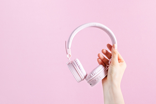 Woman hand holding pink headphones over pink background. Audio and music listening.