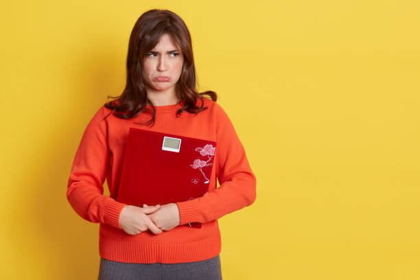 Upset young adult lady dresses casual style clothing holding floor scales in hands, looking away with pout lips, expressing sadness, bed result of loosing weight, isolated over yellow background. la joven adulta molesta viste ropa de estilo casual sosteniendo escamas de piso en las manos, mirando hacia otro lado con los labios de puchero, expresando tristeza, el resultado de la cama de perder peso, aislado sobre el fondo amarillo.