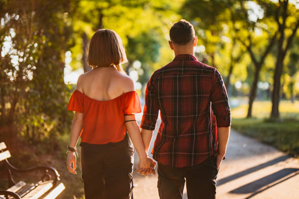 Beautiful couple in love is walking the road between the fields of sunflowers, summer sunset, bright orange light. In the background is a forest and rustic wooden houses. Concept of travel by car, eco-tourism, summer vacation, happy life, recreation in the countryside, nature beauty, gardening. Blurred background, selective focus, copy space.