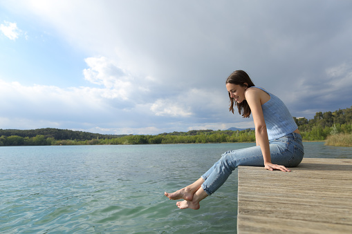 Full body profile of a happy woman resting sitting in a pier looking at lake